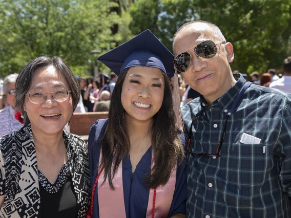 A smiling young woman in graduation cap and gown stands between her parents.