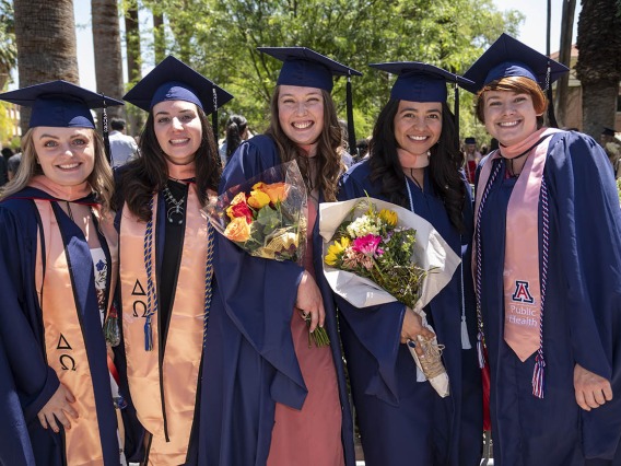 Five young women in graduation regalia stand side-by-side outside smiling, two are holding flowers. 