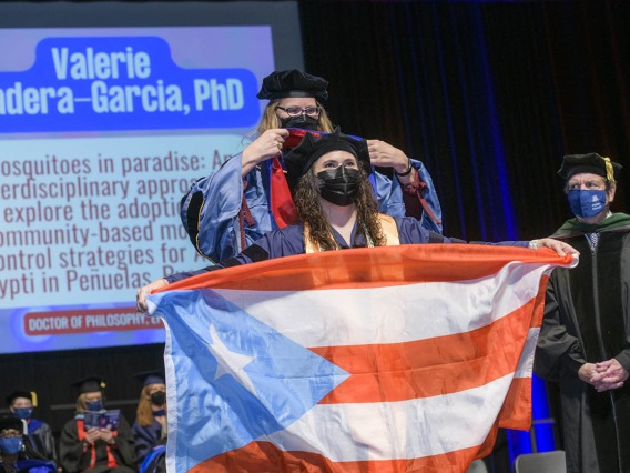 Valerie Madera-Garcia, PhD, holds the Puerto Rican flag as she is hooded during the 2022 Mel and Enid Zuckerman College of Public Health spring convocation for earning a doctorate in epidemiology.