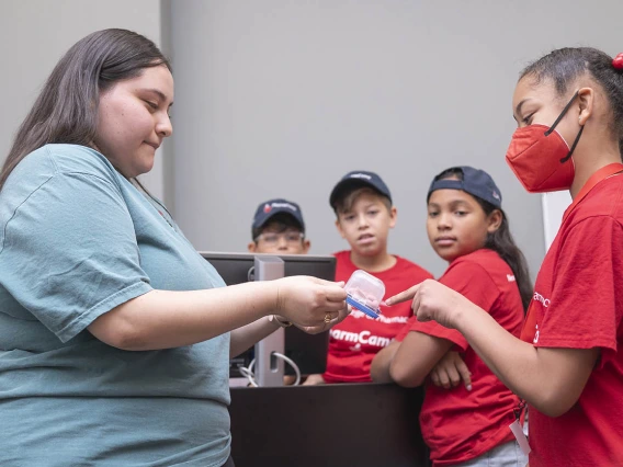 A young woman in blue shirt with long brown hair holds out a plastic container to a girl who is pointing at the container.