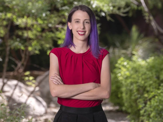 Young woman with brown and purple hair stands smiling with her arms folded. She is wearing a white short-sleeved blouse.