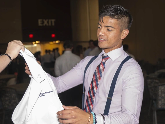 A young man with short dark hair and light brown skin wearing a button-up shirt, tie and suspenders reaches for a white coat. 