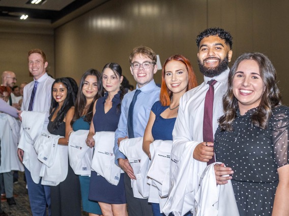 Several medical school students stand in a line smiling with white coats over their arms. 