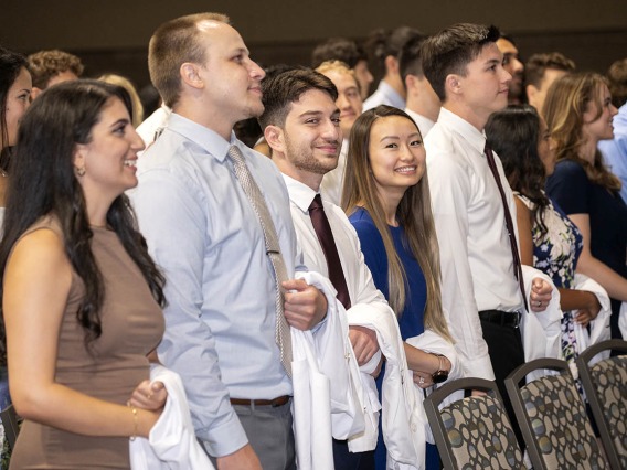 Many medical school students with white coats over their arms stand in front of chairs preparing to go onto stage.