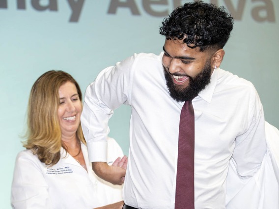 A white female doctor helps a young male medical student with brown skin and a beard put on his white coat. 