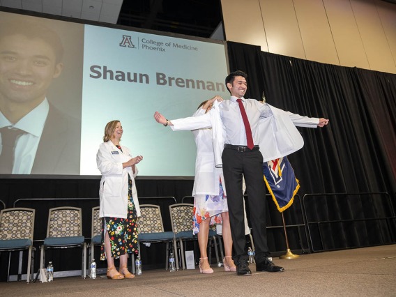 A young man stands on a stage smiling with his arms outstreatched as a faculty member helps put a white coat on him.