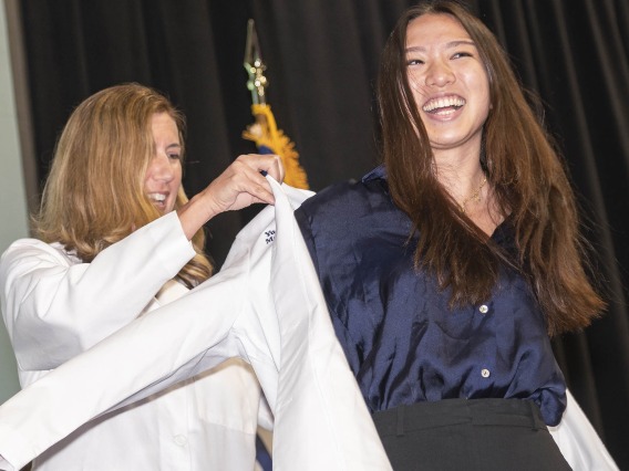 A white woman in a doctor's coat helps a young Asian woman with a big smile put on a white doctor's coat. 