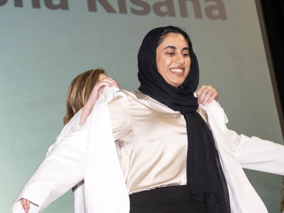 A young woman with her hair covered by a dark scarf smiles as she has her white coat placed on her. 