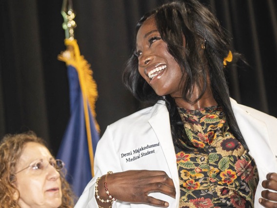 A white female doctor helps a young female medical student with dark skin and a big smile put on her white coat.