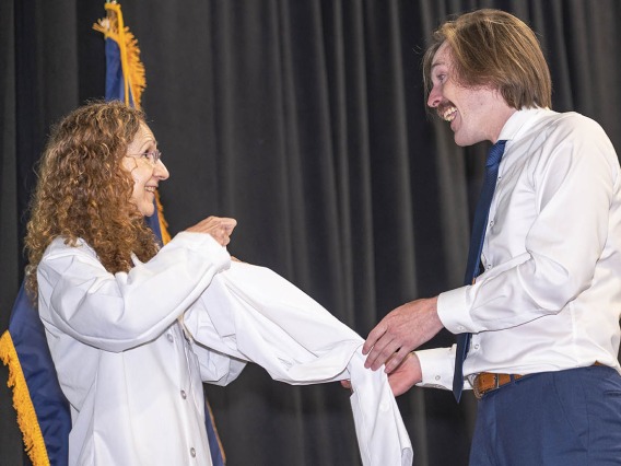 A white female doctor helps a young male medical student with light skin and a mustache put on his white coat. 
