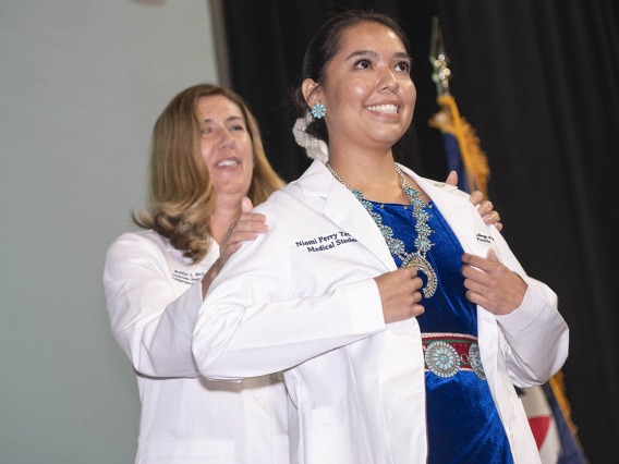 A white female doctor helps a young female medical student wearing a traditional Native American necklace and belt put on her white coat. 