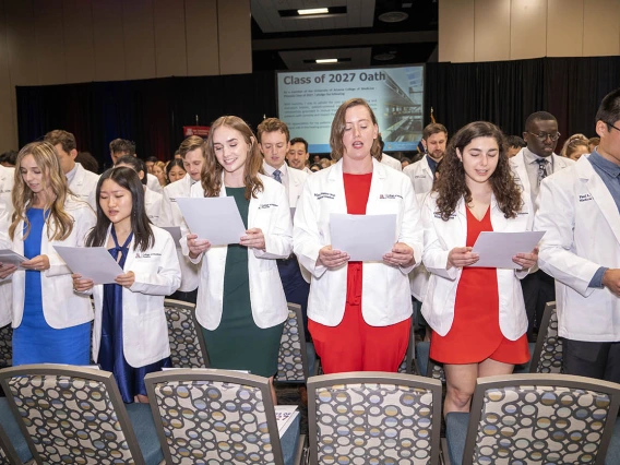 A large group of medical students wearing white coats and holding papers recites their oath.