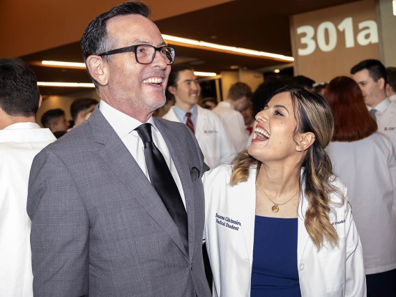 A tall white man in a suit laughs as he stands next to a shorter young woman in a white medical coat who is also laughing. 