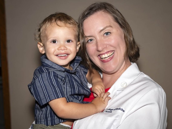 A young white woman in a medical coat smiles as she holds a little boy. Both are smiling. 
