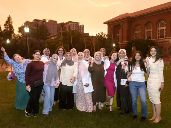 A group of about 15 women stand together, many of them medical students wearing white coats, smiling outside in the grass. 