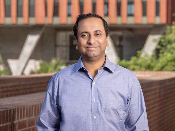 Portrait of an Indian man with short dark hair wearing a blue dress shirt in an outdoor setting. 