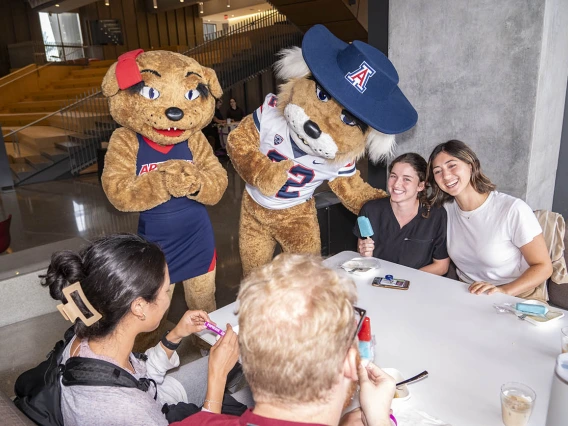 Wilma and Wilbur Wildcat mascots visit with young adults at a table while they eat. 