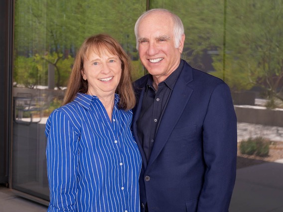 Woman with brown hair wearing a blue shirt stands with a man with grey hair wearing a black suit