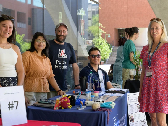 Five employees from the Zuckerman College of Public Health stand behind information tables in an outside setting. 