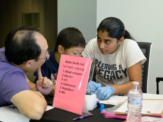 A young woman sits at a table wearing medical gloves and talks with a father and son at the Mobile Health Unit information table. 