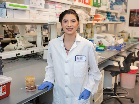 A young woman with dark hair wearing a white lab coat stands in a laboratory smiling. 