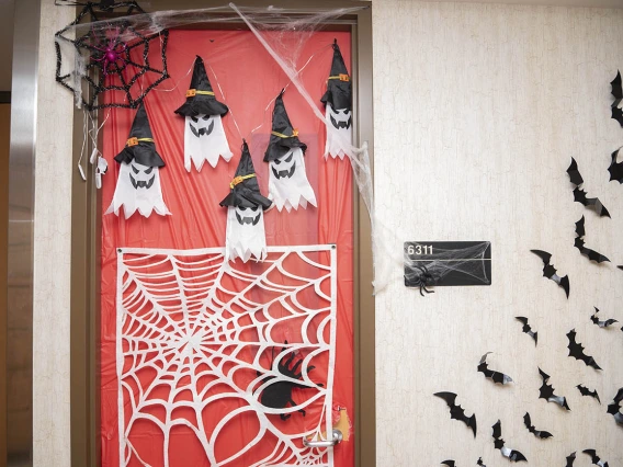 Door in a hallway covered in orange paper with several ghosts in witch's hats, a spiderweb and bats flying off on the wall to the side. 