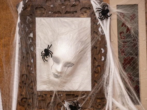 A white rectangle with a face pressing through it hangs on a door along with spiderwebs and small plastic spiders. 