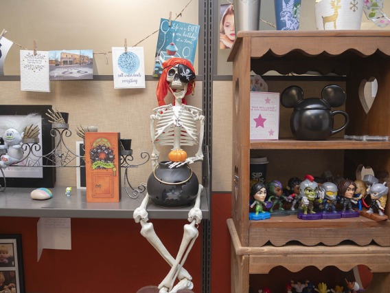 A skeleton with a pirate bandana on its head sits on a desk with a candy bowl between its legs. 