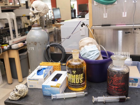 A laboratory table has larg jars labeled "Zombie Virus" along with a couple of skulls and a manikin head with a mask sitting in a bowl. 