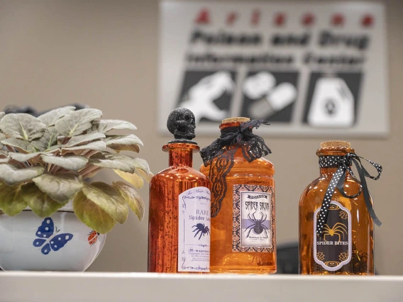 Three orange jars sit on a desk with labels that represent types of poisons. 