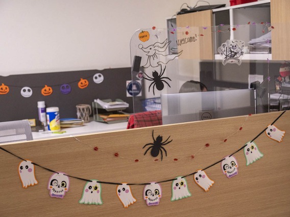 Little ghosts hang on a string across the front of a reception counter in an office. Spiders and small jack-o-lanters also decorate the area. 