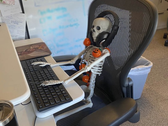 A skeleton sits in an office chair with hands on the computer keyboard and headphones on. 