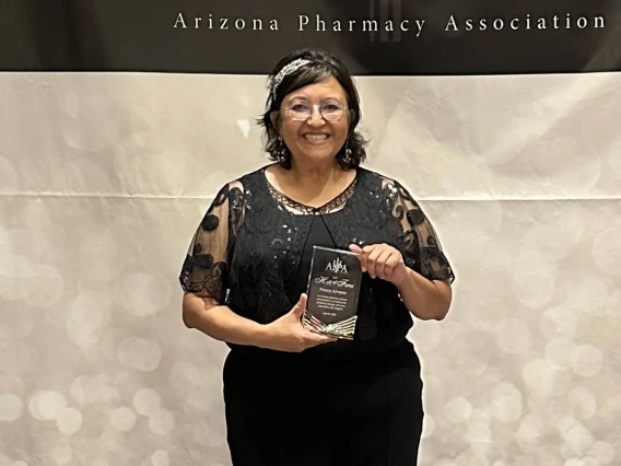 Woman with shoulder length dark hair and glasses wearing a black blouse and plants holds an award while smiling.