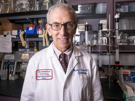 Older man with glasses and white hair stands smiling in a lab wearing a white coat.