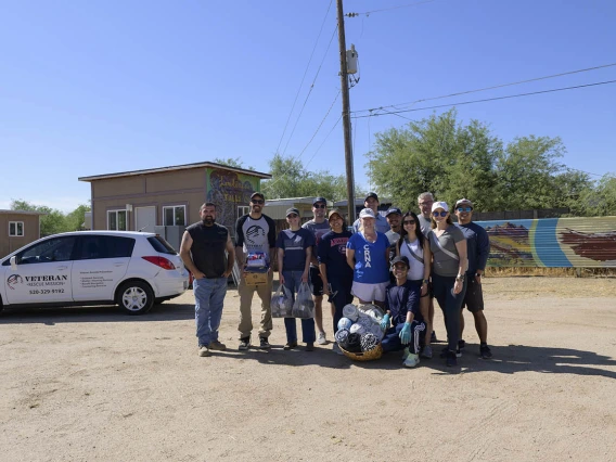 About a dozen people stand in a group in a dirt parking lot holding items they collected to donate to the Veteran Village. 