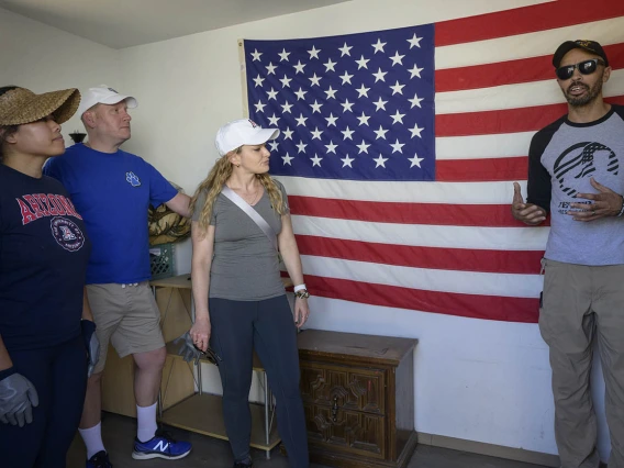 A tall man in sunglasses stands in front of an American flag talking to three volunteers from the UArizona nursing program.