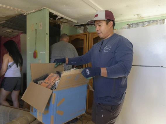 A young man wearing a baseball hat and gloves carries a large box through a rough-looking room. 