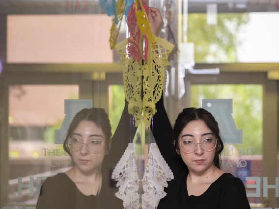 A young woman with long black hair holds up a tissue paper decoration against a window with her mirrored reflection. 