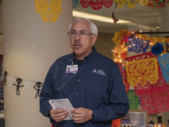Dr. Francisco Moreno, a Hispanic man with gray hair, holds a paper as he speaks in front of festivly decorated background. 