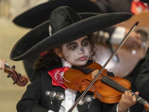 A young girl dressed in a black mariachi suit with a black sombrero hat plays a violin. Her face is painted in a traditional black and white skull design. 