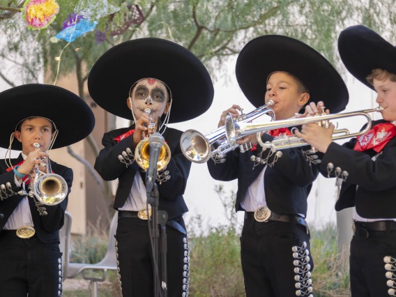 Four boys dressed in black mariachi suits with large brimmed black sombrero hats all playing trumpets.