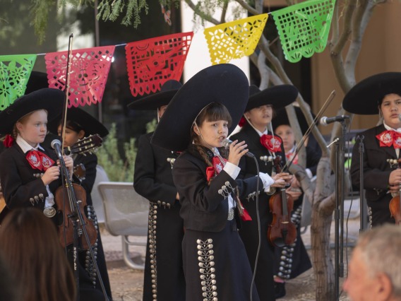 A young girl in a mariachi suit with large black sombrero sings into a microphone as severa kids in the same outfit stand around her with musical instruments. 