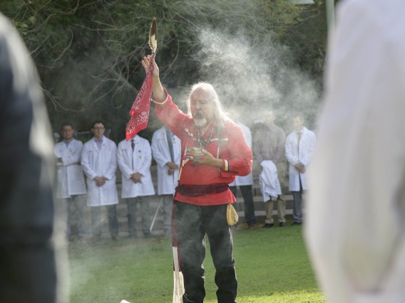 Native American man dressed in traditional tribal attire performs a ceremony surrounded by doctors dressed in white coats