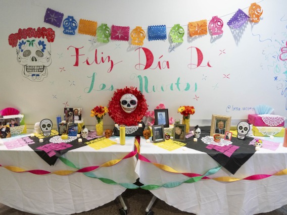 A long table is set up as a Dia de los Muertos altar with colorful streamers and skull decorations. There are photos and flowers on the table. 
