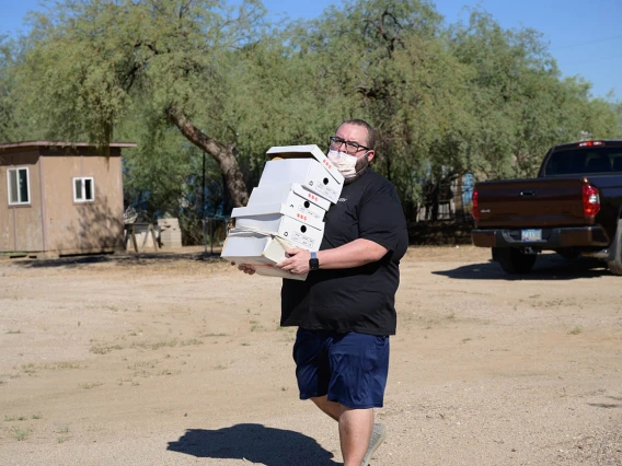 A man carries a stack of shoe boxes across a dirt parking lot. 