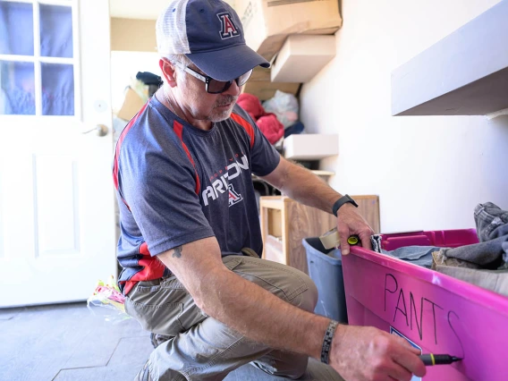 A white man squats down and writes "pants" onto the side of a pink tub full of pants. 