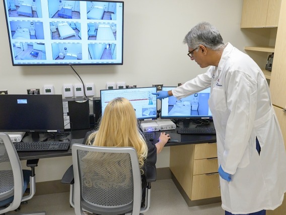 Over-the-shoulder view of a woman sitting in a chair with several monitors in front of her and a doctor standing next to her pointing at one of the screens. 