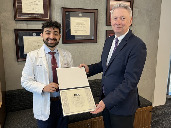 a male college dean wearing a suit presents a plaque to a male medical student wearing a white coat 
