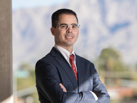 Portrait of an Hispanic man with short brown hair wearing glasses, a dark suit and a red tie. He is standing with his arms crossed and there is a mountain range behind him.