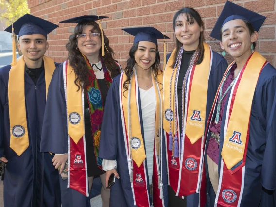 Five smiling nursing graduates in caps and gowns stand together outside. All have sashes on with the University of Arizona "A". 